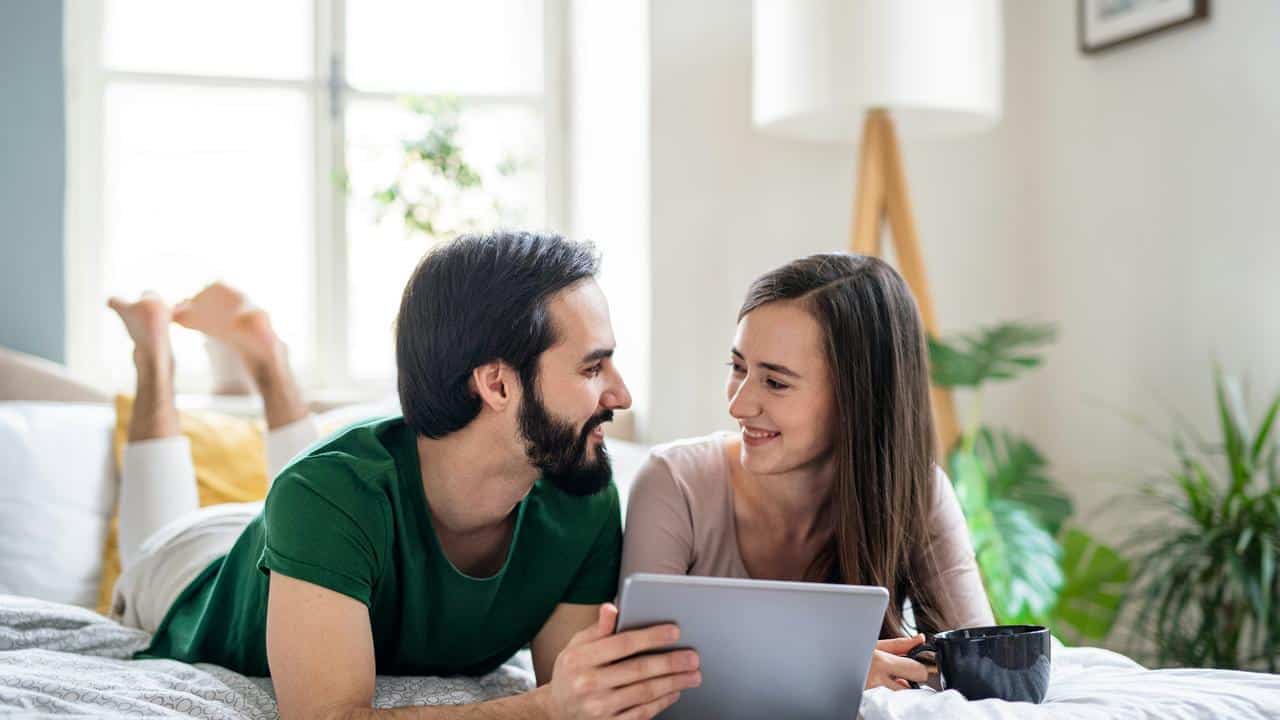 A couple smiling at each other while using a tablet in bed.