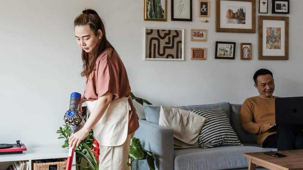 A woman vacuums a room while a man sits on a sofa using a laptop.