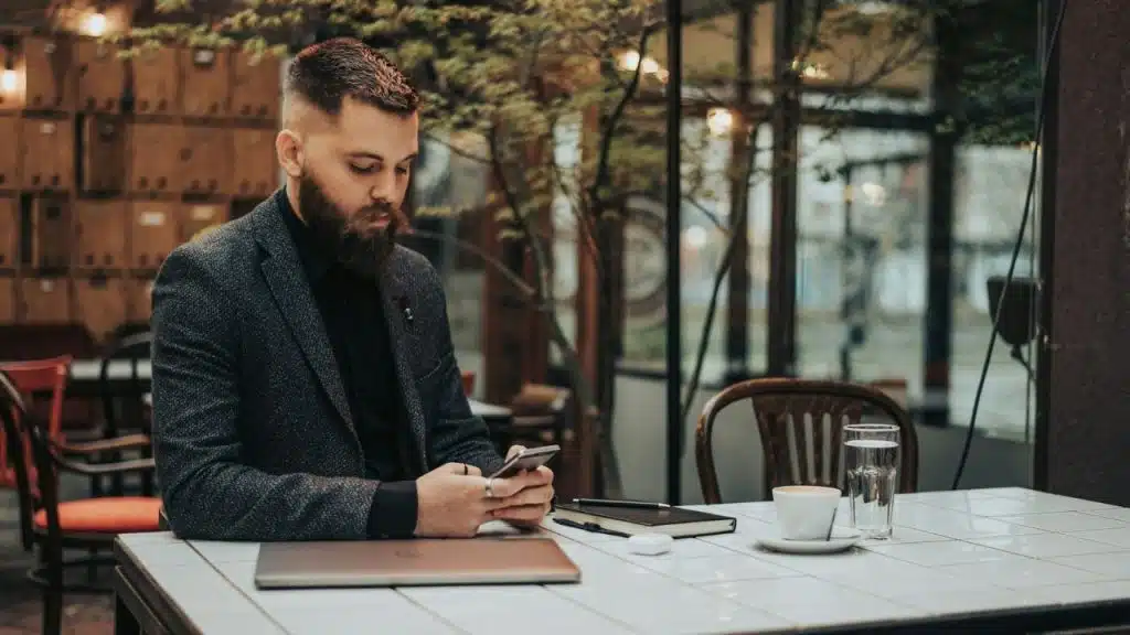 A bearded man in a blazer sits at a table and looks down at his smartphone.
