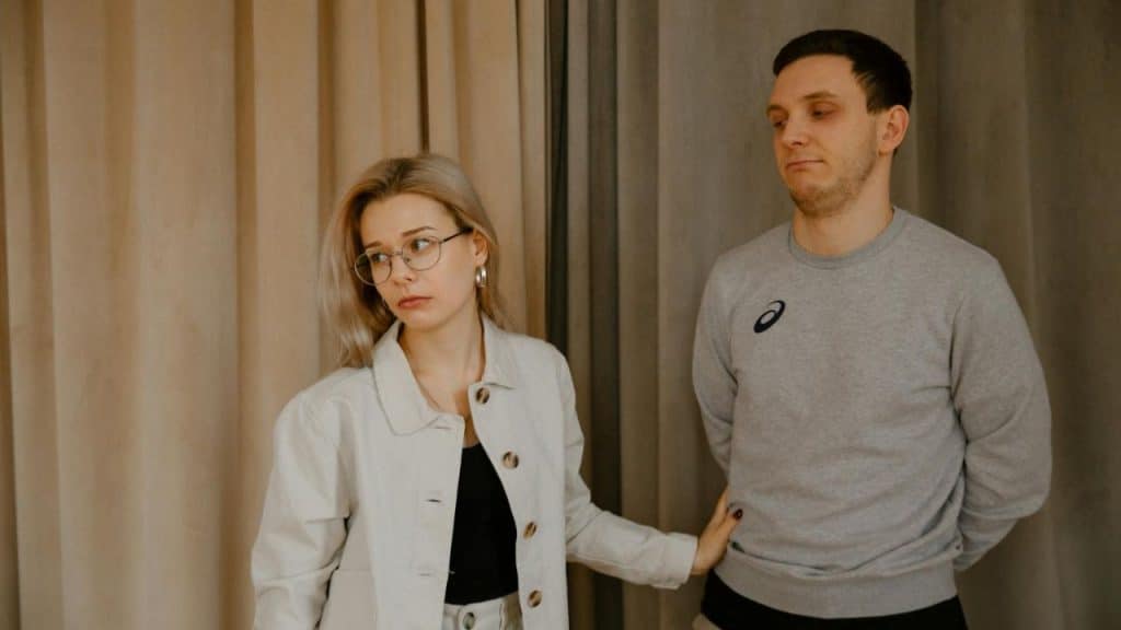A blonde woman in glasses and a man in a grey sweatshirt stand before curtains.