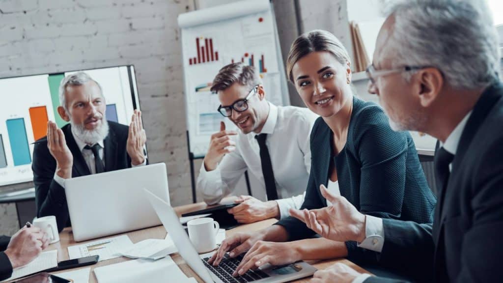 Four professionals sit at a conference table with laptops, gesturing during a group discussion.