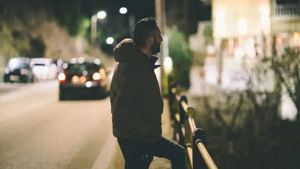 A man in a jacket leans against a metal railing at night next to a road.