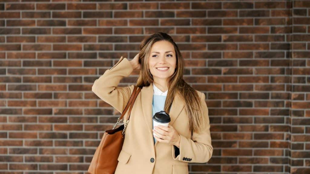 A smiling woman holding a takeaway coffee while standing against a brick wall.