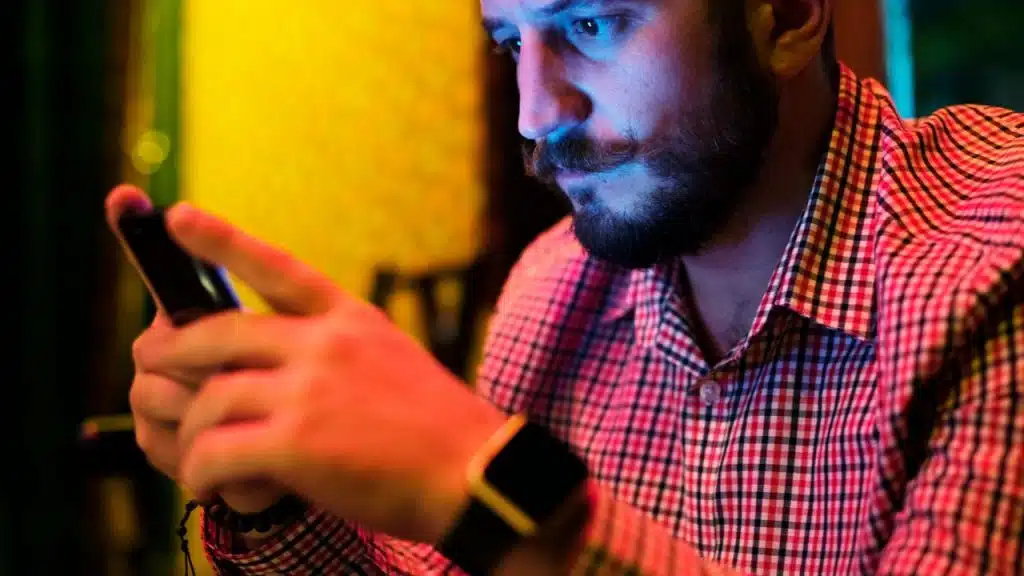 A man with a mustache looks at a glowing smartphone screen in dark, colorful lighting.