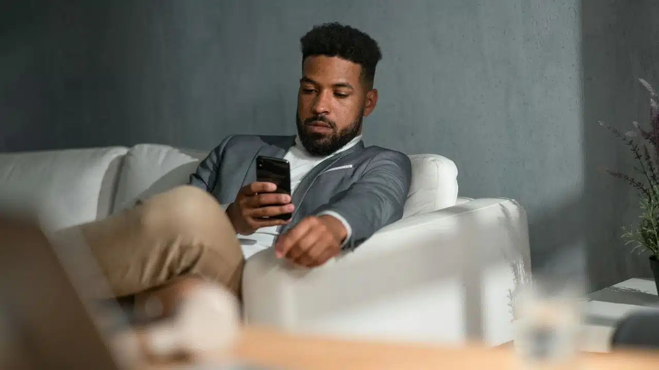 A man in a grey blazer sits on a white sofa while looking at a smartphone.