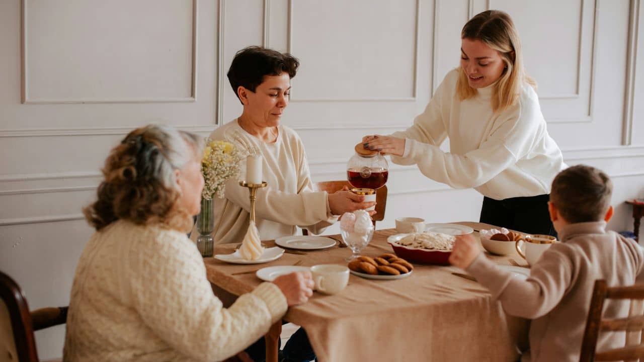 A woman pours tea for guests seated around a dining table with cookies and snacks.