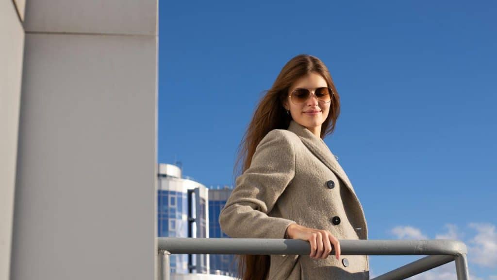 A woman in a beige coat and sunglasses leaning on a railing against a blue sky.