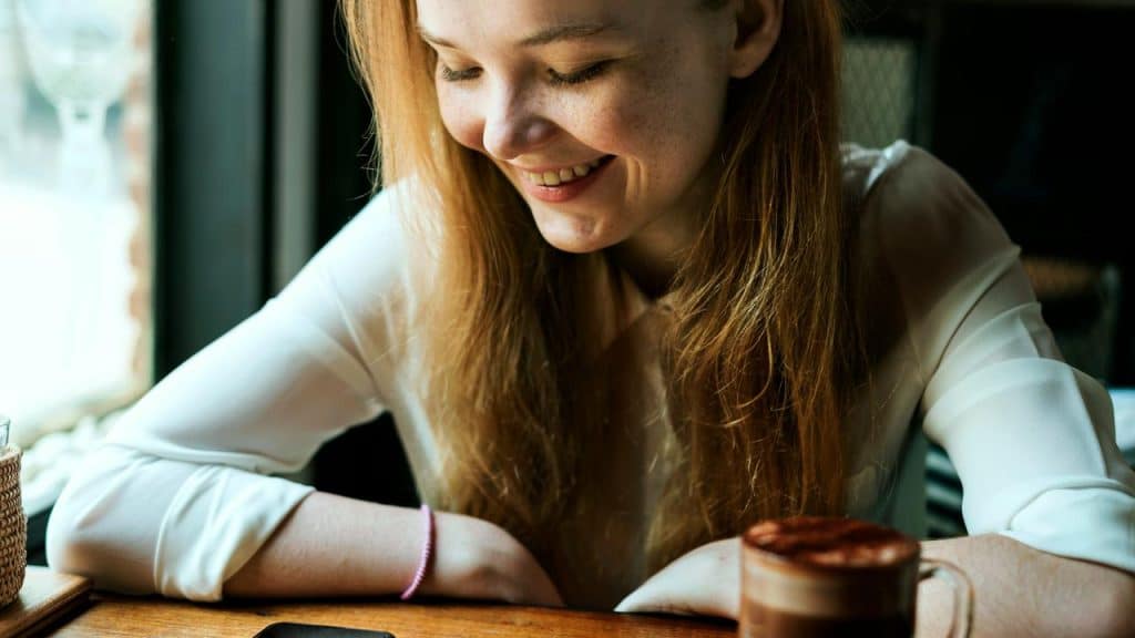 A smiling woman with freckles sitting at a café table, looking down beside a cup of coffee.