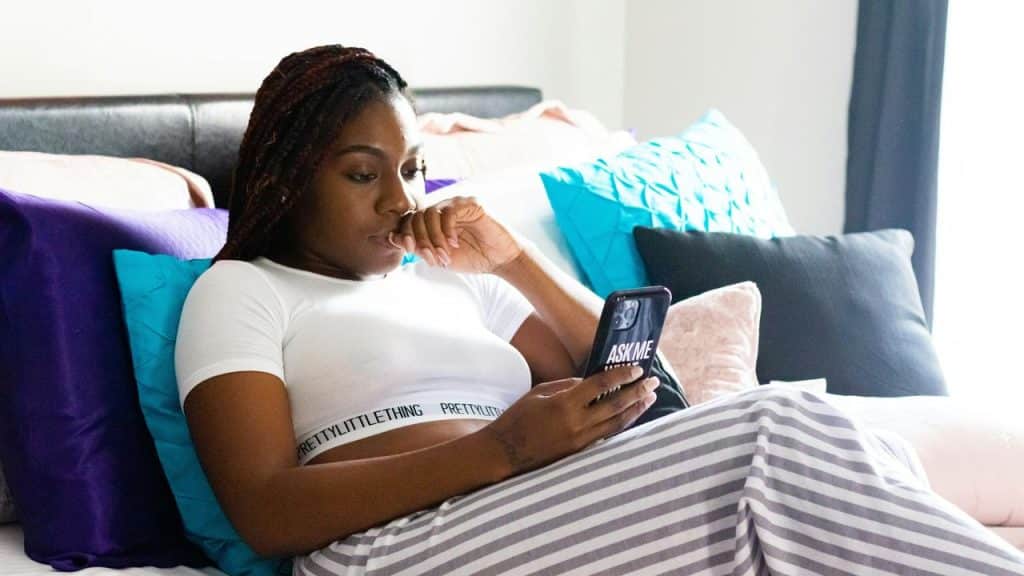 A woman lying on a bed, thoughtfully looking at her smartphone.