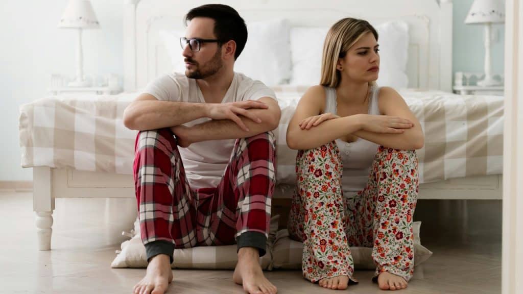 A couple sitting on the floor while leaning against a bed and looking in opposite directions and refusing to talk.