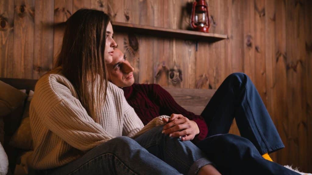 A couple sits closely together against a wooden wall while holding hands and looking away.