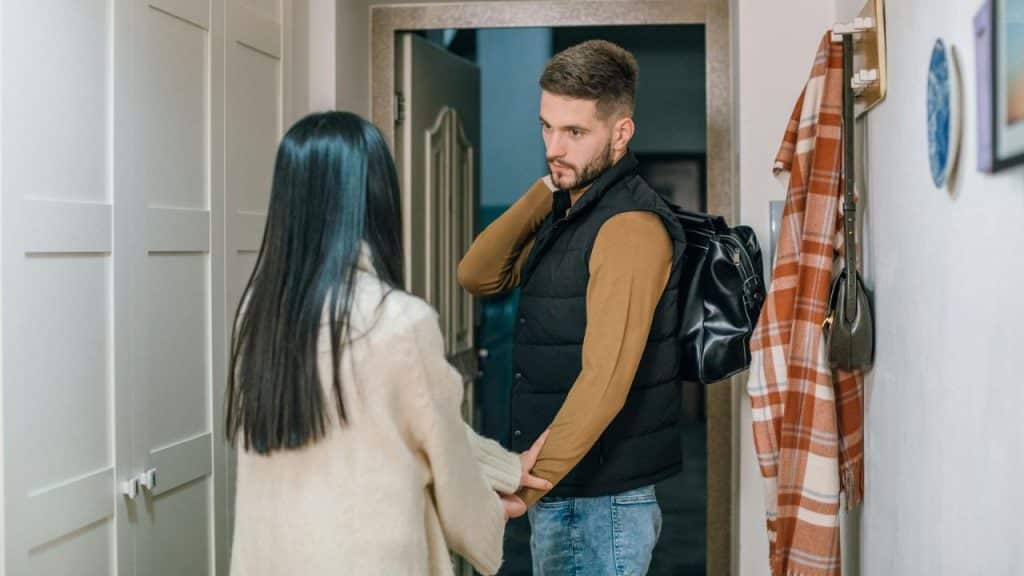 A man carrying a black leather bag looks back at a woman in a hallway.