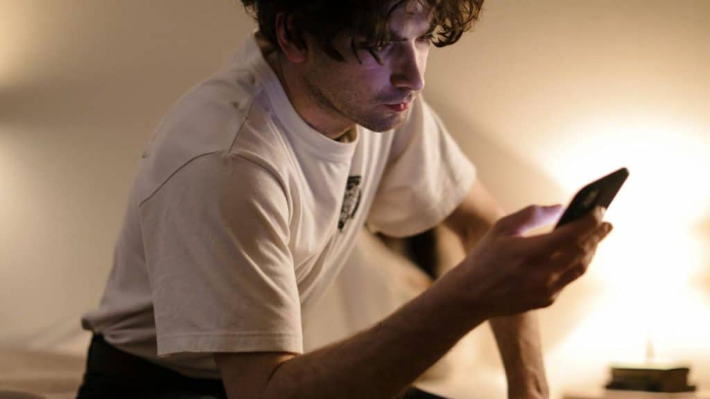 A man in a white t-shirt looks at a glowing smartphone in a dark room.