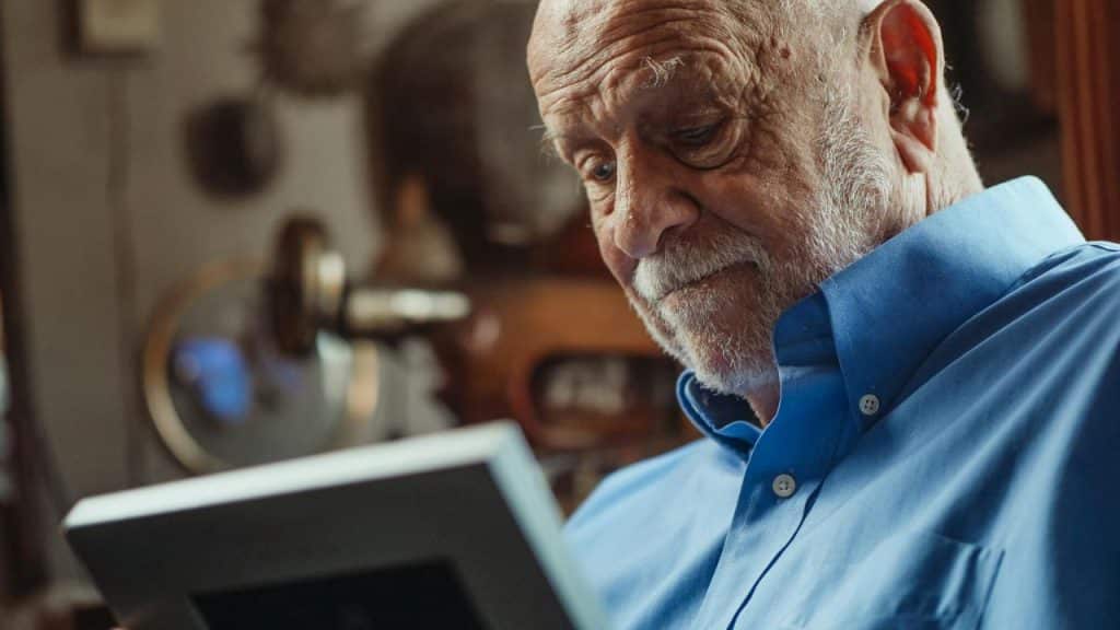 An older man with a white beard and blue shirt looks down at a framed picture.