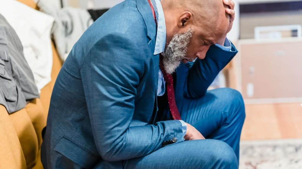 A man in a blue suit sits with his head down and hand on his forehead.