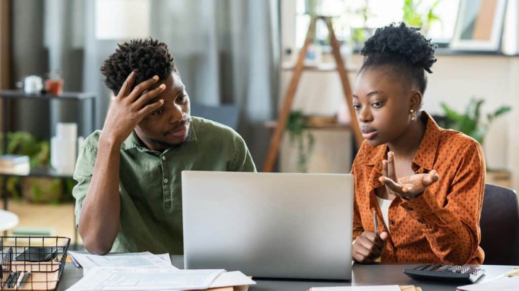 A man and woman sit at a desk with a laptop, looking at documents and gesturing.