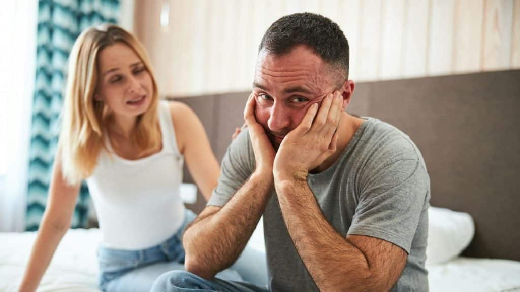 A man sits with hands on his face while a woman speaks tentatively behind him.