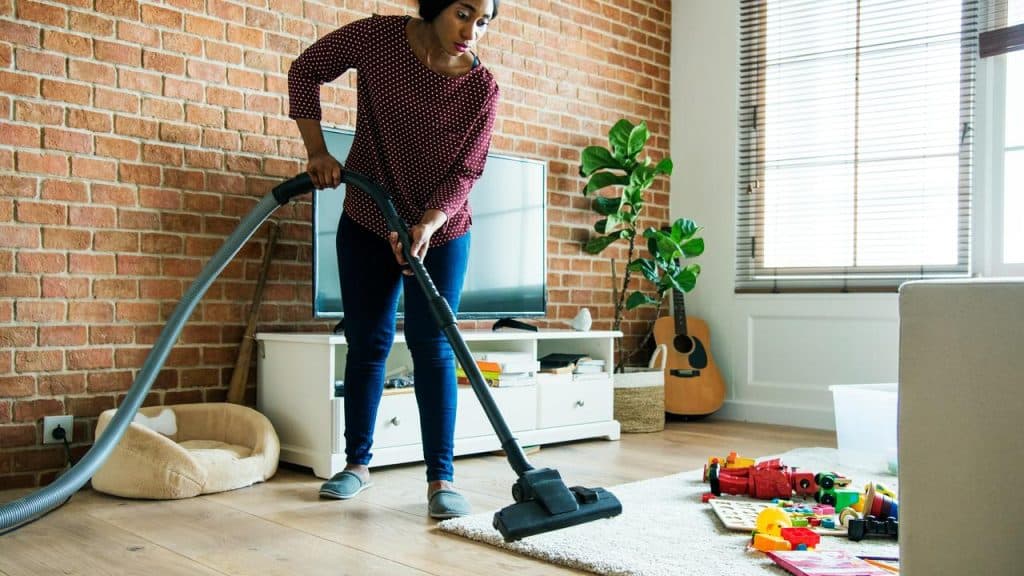 A woman vacuuming a living room floor near scattered children’s toys.