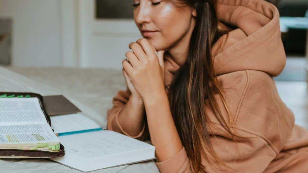 A woman with clasped hands reflecting beside open books on a bed.
