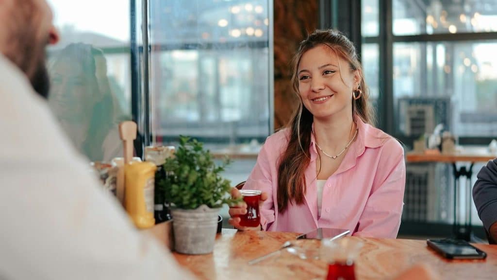 A smiling woman in a pink shirt holding a glass of tea at a café table.