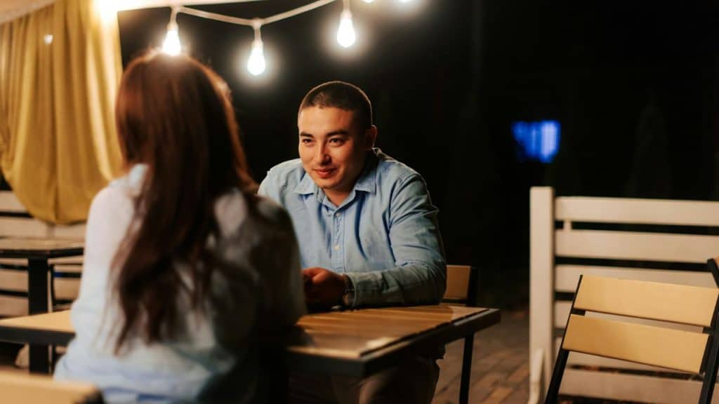 A man and woman sit at an outdoor table at night under warm string lights.