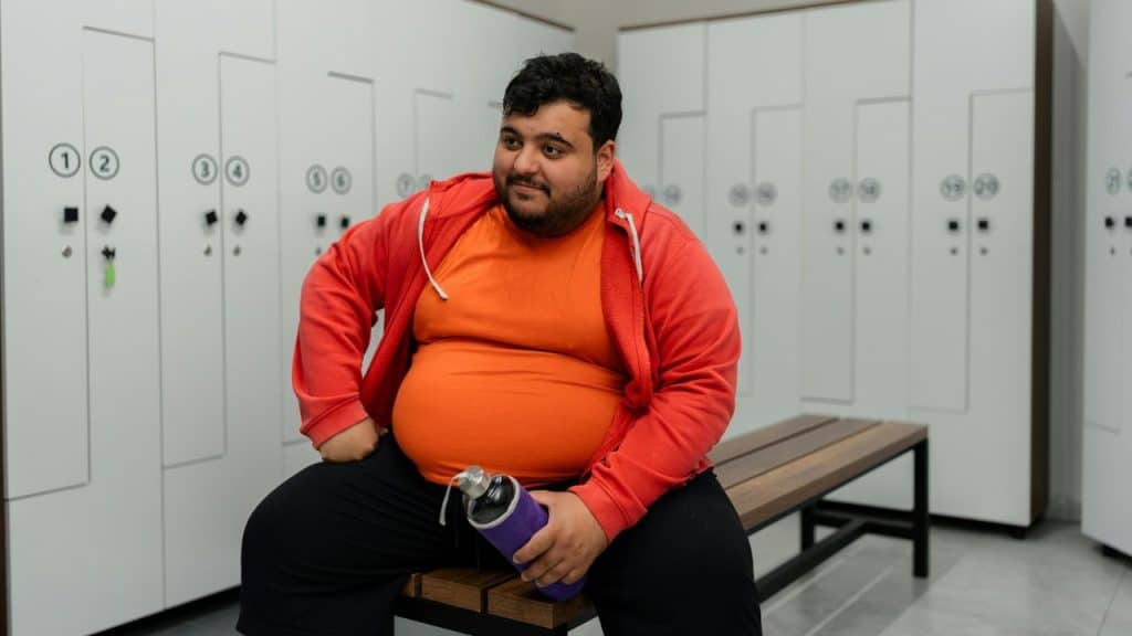A man in an orange shirt and red jacket sits in a locker room with a bottle.