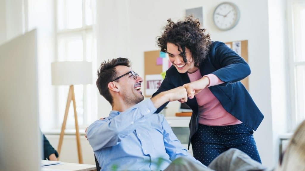 A man and woman laugh while performing a fist bump in a bright office setting.