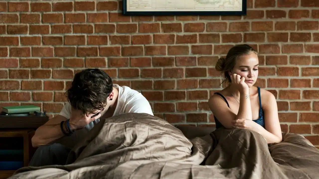 A man and woman sit apart in bed with a brick wall in the background.