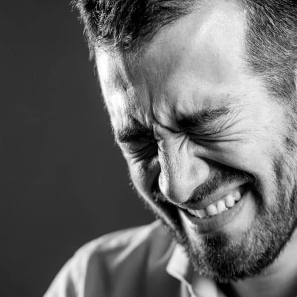 Black and white close-up of a bearded man with eyes tightly shut and teeth bared.