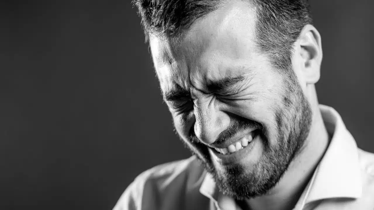 Black and white close-up of a bearded man with eyes tightly shut and teeth bared.