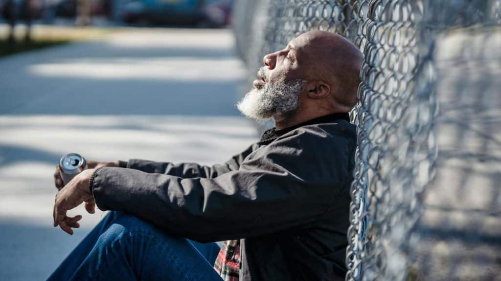 A bald man with a grey beard leans against a chain-link fence holding a silver can.