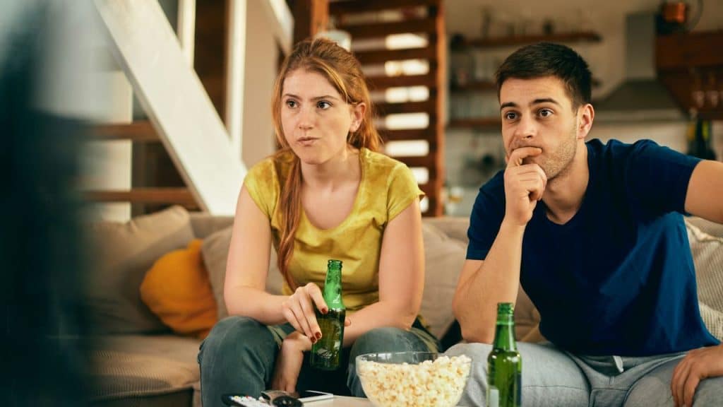 A man and woman sit on a sofa with beer bottles and a bowl of popcorn.