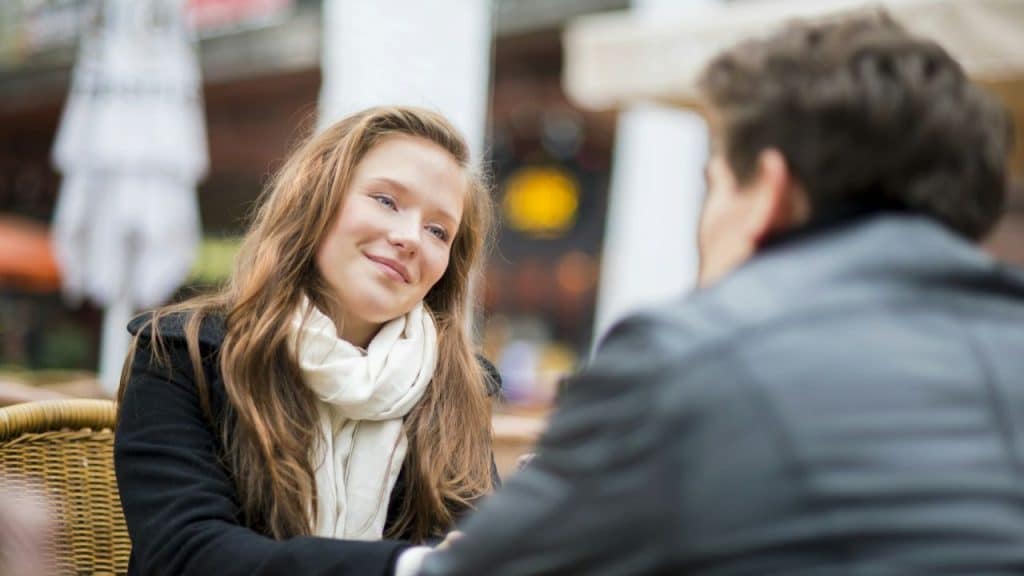A woman with long light-brown hair and a white scarf smiles at a man outdoors.