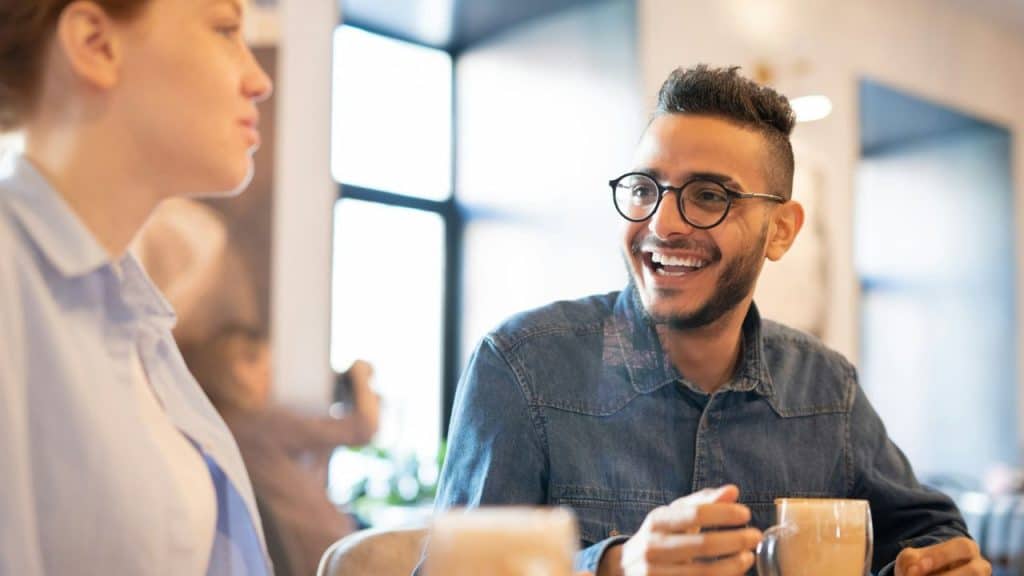 A smiling man with glasses talks to a woman at a table in a cafe.