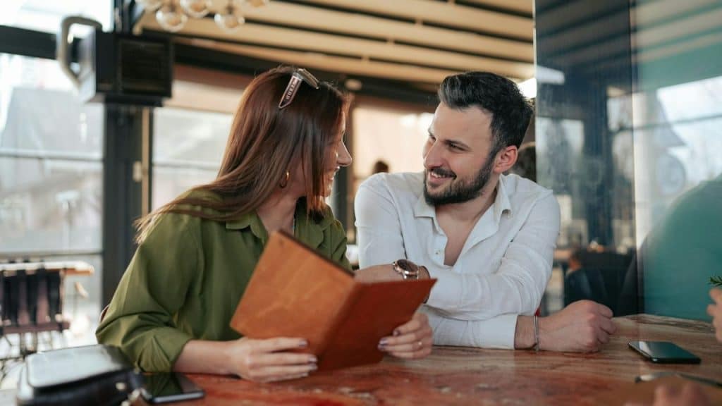 A man and woman sit at a wooden table smiling while looking at a menu together.