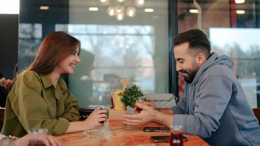 A woman and a man sit at a wooden table, smiling and looking at each other.