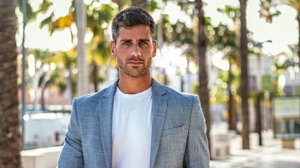 A man in a light blue blazer and white t-shirt stands outdoors with palm trees.