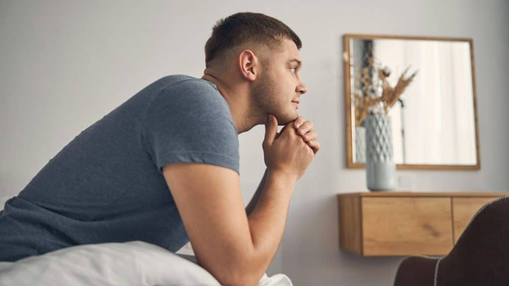 A man sits on a bed with his chin resting on his interlaced hands, looking away.