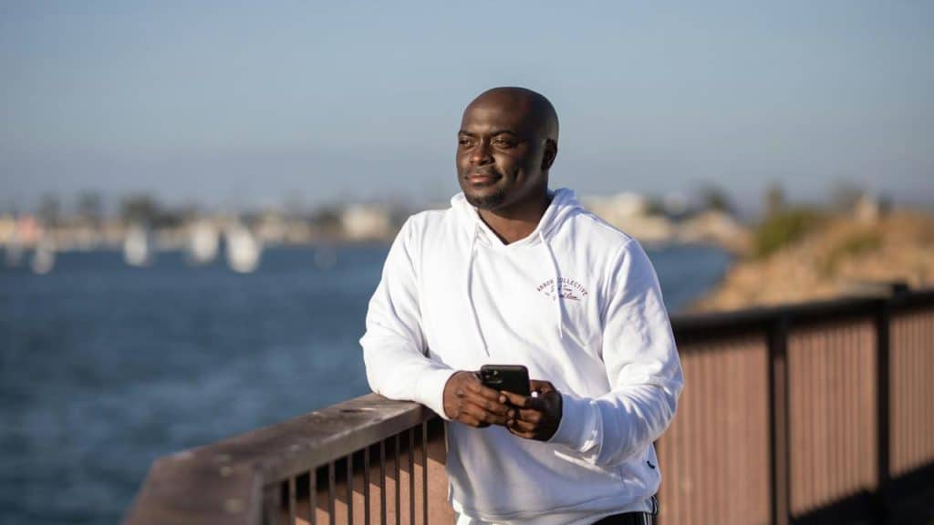A man in a white hoodie holding a phone while standing by a waterfront railing.