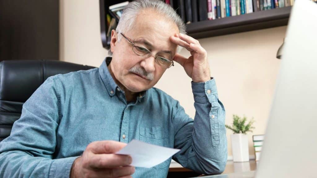 An older man with glasses sits at a desk, looking down at a piece of paper.
