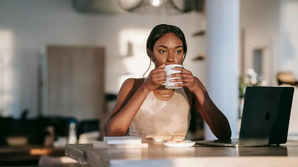 A woman sipping coffee while sitting at a table with a laptop in a bright, modern interior.