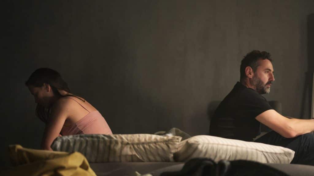 A man and a woman sit back-to-back on a bed in a dimly lit room.