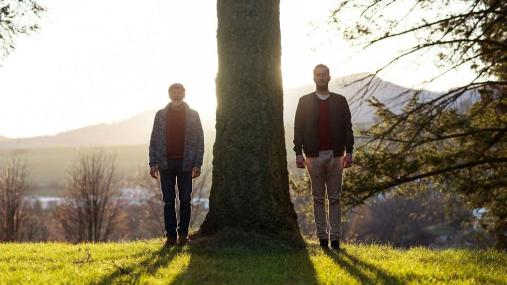 Two men stand on opposite sides of a large tree trunk in a grassy field.