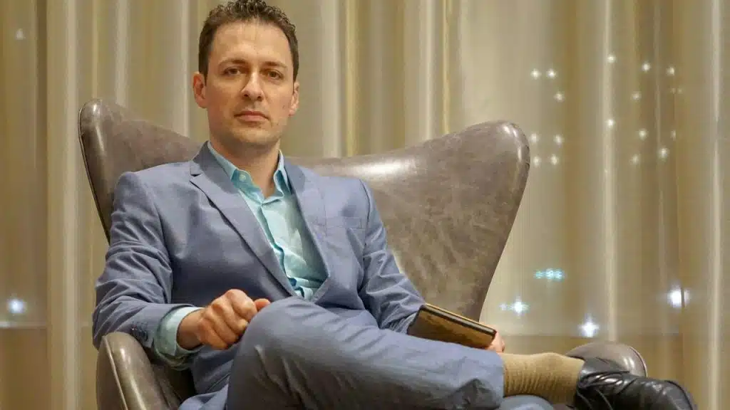 A man in a blue suit sits in a chair holding a book with curtains behind.
