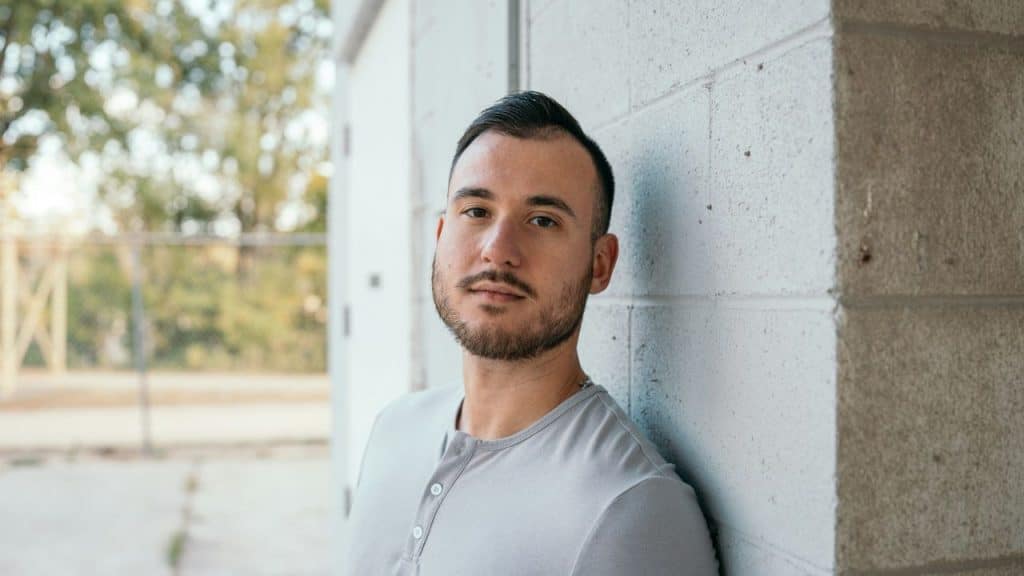 A man in a gray shirt stands outside, leaning his shoulder against a white brick wall.