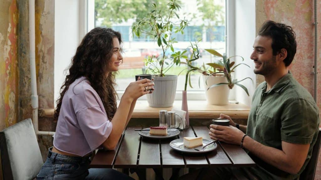 A man and woman sit at a cafe table with coffee and cheesecake, smiling together.