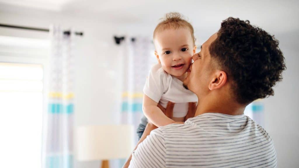 A father holding and kissing his smiling baby in a bright, softly lit room.