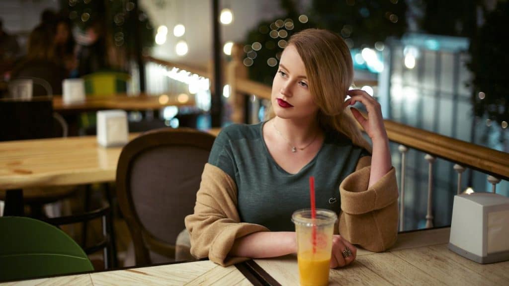 A blonde woman with red lipstick sits at a table in a dimly lit restaurant, holding her hair and looking away