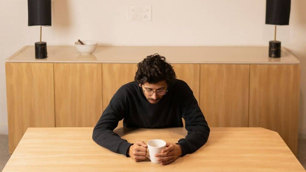 A man with glasses sits at a wooden table looking down at a white mug.