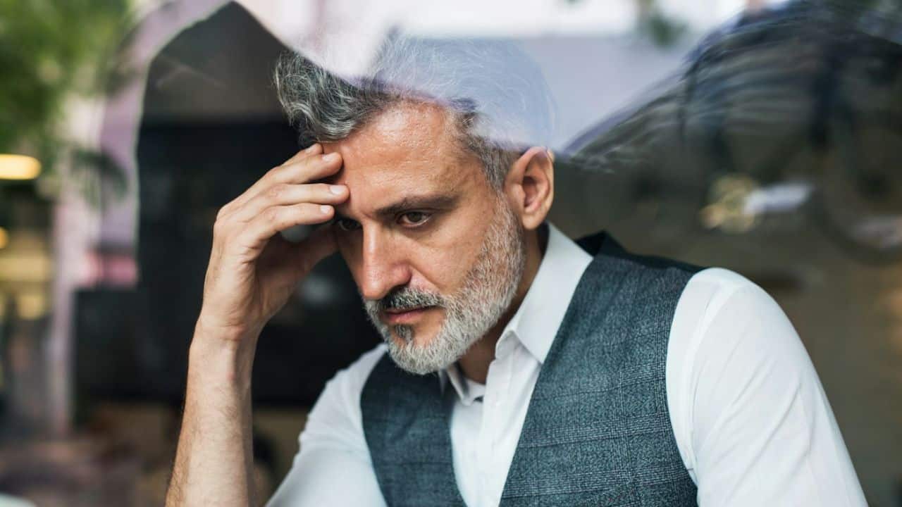 An older man with graying hair sits by a window, looking down while resting his forehead on his hand.